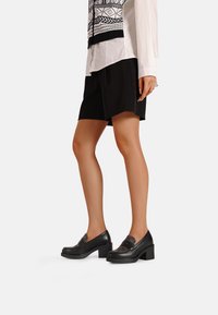Scarpe in pelle nera con tacco a blocco e accenti in tessuto decorato sulla parte anteriore, abbinate a pantaloni corti neri e una blusa bianca a fantasia.