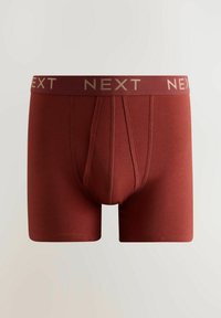 Raudonos bokso stiliaus apatinės kelnaitės iš minkštos medžiagos, su plačiu bordo spalvos liemeniu, ant kurio auksinėmis raidėmis užrašyta „NEXT“, ir prigludusiu dizainu.