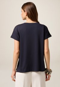 Camisa de manga corta de color azul marino, con cuello redondo, corte holgado, aberturas laterales, combinada con una pulsera estampada, fabricada en algodón suave.