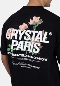 Černé bavlněné tričko s bílým textem a květinovým potiskem na zadní straně, které zobrazuje název značky "CRYSTAL PARIS" a slogan.