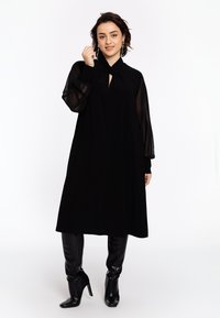 Robe noire avec de longues manches en voile, détail de col en forme de clé, et une coupe décontractée. Associée à des bottes noires brillantes à talons, montantes jusqu'au genou.