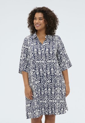 Kobieta z kręconymi włosami, ubrana w granatowo-białą sukienkę do kolan z guziczkami, uśmiechająca się i patrząca na bok.