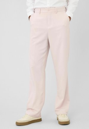 Personne portant un pantalon droit rose clair avec poches latérales et des baskets blanches avec semelles marron, debout devant un fond gris.