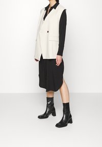 Débardeur sans manches crème avec un col en V, des boutons décoratifs et des poches sur les côtés, superposé à une robe noire à boutons. Bottines noires à bout carré.