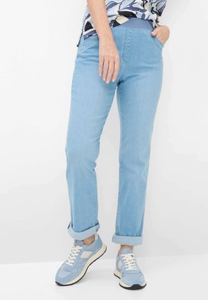 Person iført lyseblå bukser med opkantsede ben og matchende lyseblå sneakers, stående med den ene hånd i lommen mod en hvid baggrund.