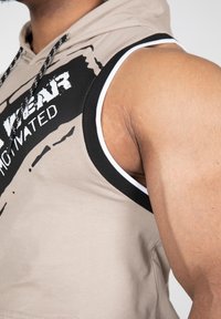 Lichtbeige mouwloze hoodie met zwarte grafische tekst, zwart-wit afwerking op de armgaten en een gestructureerde stof, die de gespierde armdetails toont.