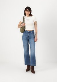 Valkoinen slim-fit T-paita logolla, korkeavyötäröiset siniset farkkuleggingsit, ruskeat nilkkurit ja pieni oliivinvihreä olkalaukku.