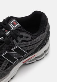 Musta urheilukenkä, jossa on verkkomateriaali yläosassa, teksturoidut pinnoitteet, heijastavat yksityiskohdat ja vaahtomuovista valmistettu valkoisharmaa pohja. Sivulla on logo.