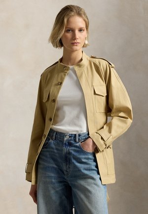 Woman wearing beige buttoned jacket with pockets over white top and blue jeans, hand in pocket, against plain neutral background.