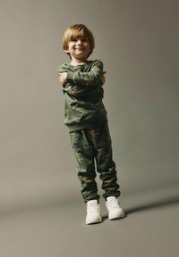 Sudadera verde y pantalones a juego, ambos con coloridos parches temáticos de aventura; combinados con zapatillas blancas. Tejido suave y diseño informal.
