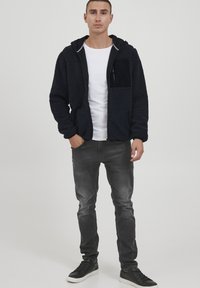 Black fleece-lined zip-up hoodie with a front pocket, paired with gray slim-fit jeans and black sneakers. Simple design, casual fit.