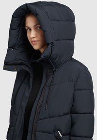 Marineblaue Pufferjacke mit großer Kapuze, gestepptem Design, Frontreißverschluss, Kordelzügen und Logodetail an der Seite; glatte Textur.