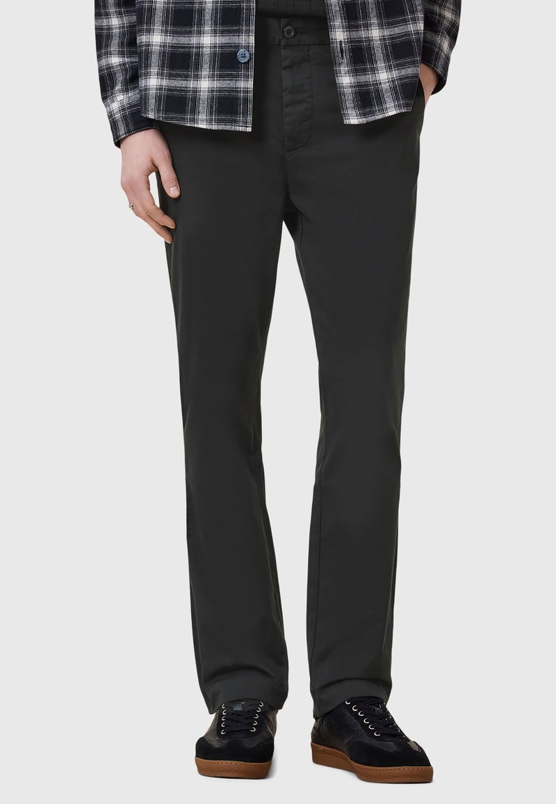 Muž v černých rovně střižených kalhotech, černobíle kostkované košili a černých teniskách s hnědými podrážkami, stojící s rukama v kapsách.