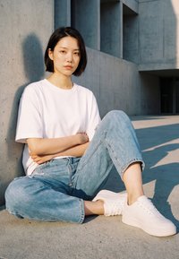 Nuori nainen valkoisessa t-paidassa, vaaleansinisissä farkuissa ja valkoisissa lenkkareissa istuu risti-istunnassa betonilattialla harmaata seinää vasten auringonpaisteessa.