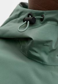 Lysegrønn jakke med høy krage, utstyrt med svart knapp og justerbar snor. Glatt, vanntett stoff, med minimale sømdetaljer.