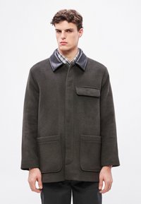 Σκούρο πράσινο μάλλινο παλτό με μαύρο δερμάτινο γιακά, μία τσέπη στον στήθος και δύο μπροστινές τσέπες· ημικαλομολογιακή γραμμή και απαλή υφή.