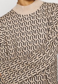 Maglione lavorato a maglia beige con un motivo geometrico nero. Presenta un alto colletto a coste e maniche lunghe e morbide. Tessuto morbido, design casual.