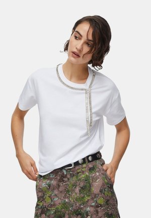 Donna che indossa una maglietta bianca con dettaglio a catena d'argento intorno al collo e pantaloni a fantasie floreali con una cintura nera.