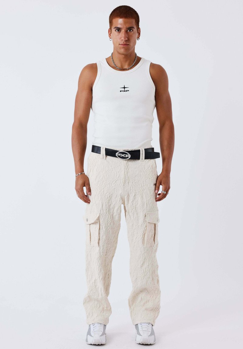 Jeune homme portant un débardeur blanc, un pantalon cargo crème texturé, une ceinture noire avec une boucle argentée, des bijoux en argent et des baskets blanches, debout devant un fond blanc.