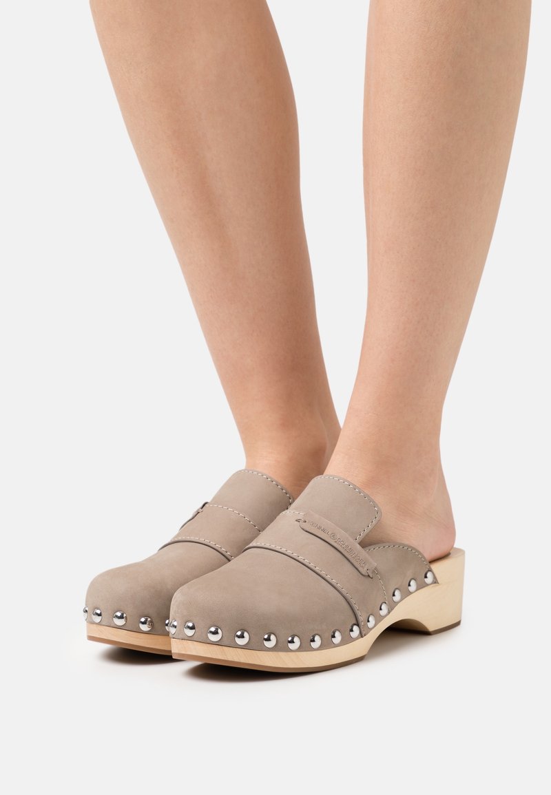 Pieds portant des sabots en cuir taupe avec des clous argentés le long des bords et des petits talons blocs en bois sur fond blanc.