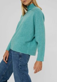 Pull turquoise côtelé avec un col montant et des manches longues, associé à un jean en denim bleu. Le tissu semble doux et texturé.