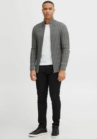Grauer gestrickter Reißverschluss-Pullover mit strukturiertem Muster, kombiniert mit einem weißen Hemd und schwarzen Hosen. Schwarze Sneakers runden das Outfit ab.