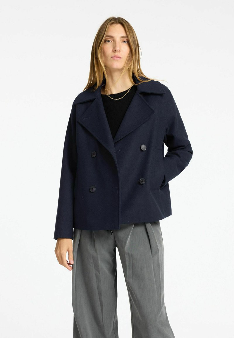 Cappotto peacoat doppiopetto blu navy con grandi bottoni, ampio colletto a punta e vestibilità rilassata. Indossato sopra una maglia nera e pantaloni plissettati grigi.