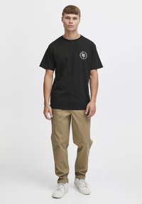 Sort bomulds t-shirt med et cirkulært hvidt graphic på brystet, parret med beige bukser og hvide sneakers.