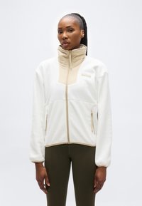 Γυναίκα που φοράει λευκό και μπεζ φλις μπουφάν με φερμουάρ και ψηλό γιακά, καθώς και ελαιί πράσινο κολάν, στέκεται μπροστά σε απλό φωτεινό φόντο.