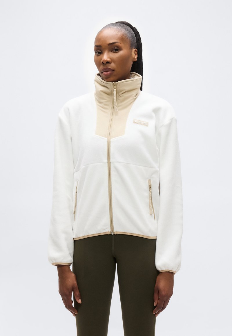 Γυναίκα που φοράει λευκό και μπεζ φλις μπουφάν με φερμουάρ και ψηλό γιακά, καθώς και ελαιί πράσινο κολάν, στέκεται μπροστά σε απλό φωτεινό φόντο.