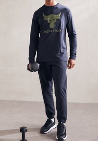 Mann in schwarzem Sportoutfit, der mit einer Hand eine Kurzhantel hält, während er neben einer weiteren Kurzhantel auf dem Boden steht.