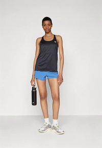 Czarny sportowy top na ramiączkach i niebieskie szorty. Modelka trzyma czarną butelkę na wodę. Buty mają szarobiały wzór z kontrastującymi detalami.