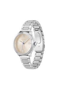 Reloj de acero inoxidable plateado con una esfera redonda, dial texturizado en dorado claro, manecillas azules y un diseño de pulsera con cierre. Logo de Lacoste presente.