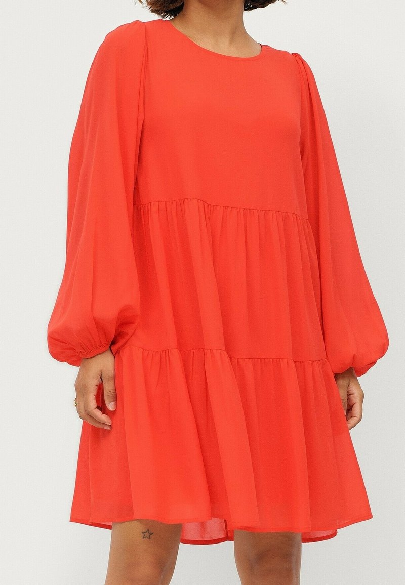 Femme portant une robe ample, rouge vif, à manches longues et à étages, avec des manches bouffantes et un col rond, debout devant un fond uni.