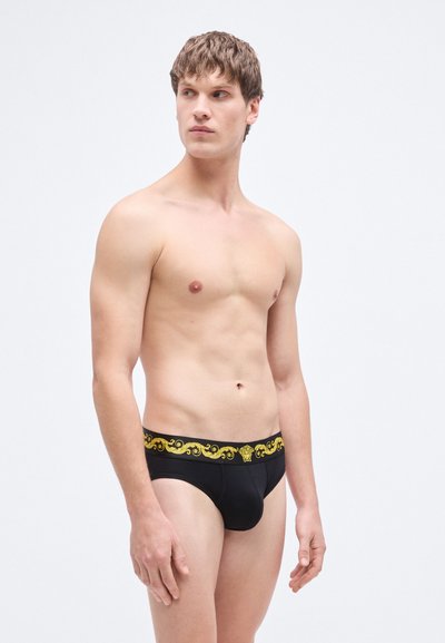 Caleçon noir avec une ceinture dorée à motifs ornés. Le tissu semble lisse et épouse parfaitement la silhouette d'un modèle masculin.