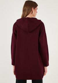 Bordo didelis džemperis su tekstūru, gobtuvu ir ilgomis rankovėmis. Yra paprasto dizaino, be matomų detalių ar akcentų.