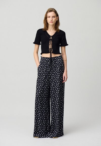 Top de punto negro recortado con un diseño de lazo en el frente, combinado con pantalones sueltos negros que presentan un patrón floral blanco.