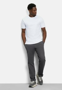 Weißes T-Shirt mit kurzen Ärmeln, kombiniert mit dunkelgrauen Hosen und schwarzen Sneakers. Das T-Shirt hat einen Rundhalsausschnitt und eine glatte Textur.