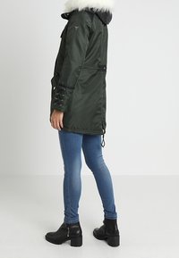 Parka verde con cappuccio foderato in pelliccia, dotata di chiusura con zip, cordini regolabili e dettagli con lacci sulle maniche, indossata sopra jeans blu.