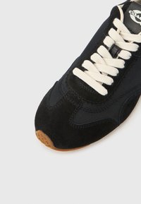 Μαύρο σουέντ αθλητικό παπούτσι με λευκά κορδόνια και σόλα από καουτσούκ χρώματος καραμέλας. Διαθέτει ζιγκ ζαγκ διακοσμητικά στοιχεία και ραφές, σχεδιασμός με διπλά υλικά.