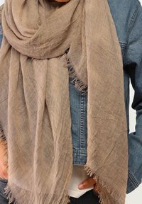 Écharpe beige texturée aux bords frangés, drapée sur une veste en jean bleue portée par une personne avec la main posée près de la taille.