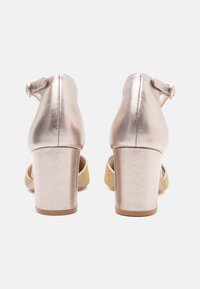 Para metalicznych sandałów w kolorze różowego złota na słupku z paseczkami wokół kostki i błyszczącym złotym paskiem na palcach, widziana z tyłu na białym tle.