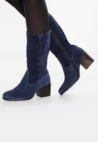 Ben i svarta tights bär marinblå mocka stövlar till medellång längd med klossklackar och synliga sidodragkedjor mot en vit bakgrund.