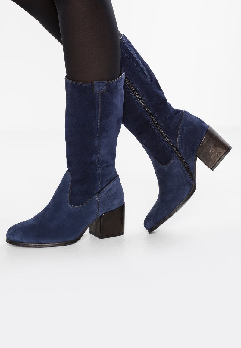 Ben i svarta tights bär marinblå mocka stövlar till medellång längd med klossklackar och synliga sidodragkedjor mot en vit bakgrund.