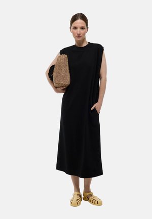 Robe noire sans manches avec des poches latérales, le tissu tombe librement. Le mannequin tient un sac tissé texturé et porte des sandales ouvertes jaunes.