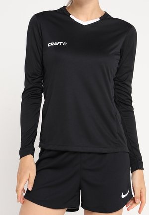 Donna che indossa una maglietta sportiva nera a maniche lunghe con il logo "Craft" e pantaloncini Nike neri, in piedi davanti a uno sfondo semplice.
