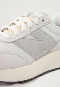 Grå sneaker med en texturerad, sprucken läderdetalj, meshnätpaneler och platta snören. Lätt sula med en svart yttersula med mönstring.