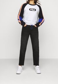 Sweatshirt blanc avec des manches noires et colorées, arborant un logo. Un pantalon noir et des sneakers blanches avec des accents texturés complètent le look.