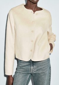Persona che indossa un cardigan di lana color crema con bottoni e jeans a vita alta di colore blu chiaro, con una mano in tasca, su uno sfondo semplice.
