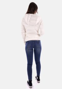 Giacca a vento bianca con cappuccio, design trapuntato e silhouette aderente, indossata con jeans blu scuro e sneakers nere.